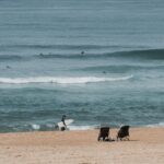 A surfer walks along the sandy beach of Hossegor, France, with waves rolling in.