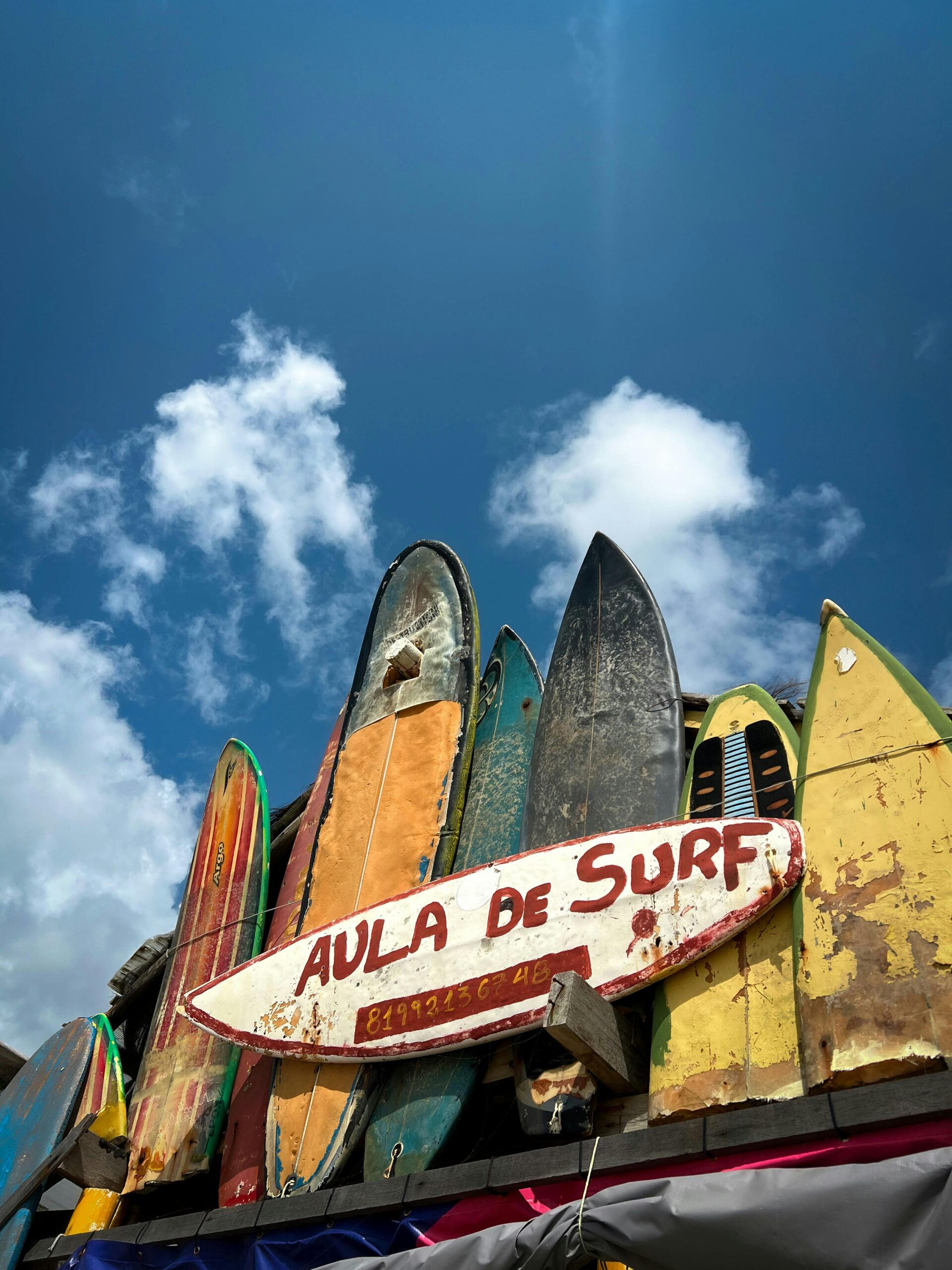 A collection of vibrant surfboards with 'Aula de Surf' sign against a bright blue sky in Brazil.